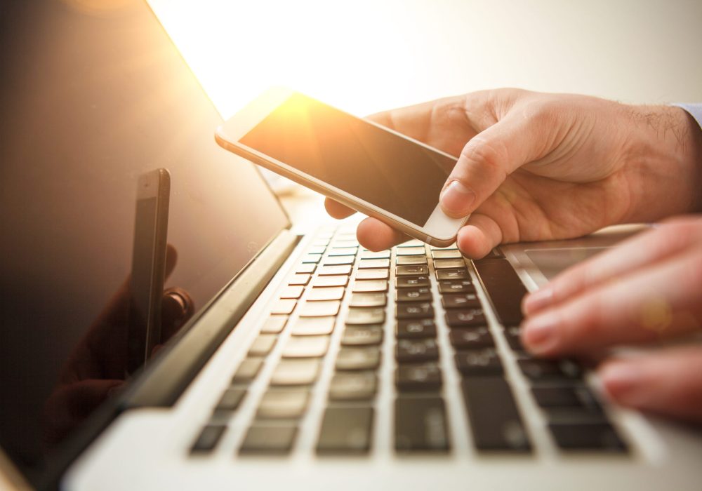 The male hand holding a phone against the background of the office and laptop